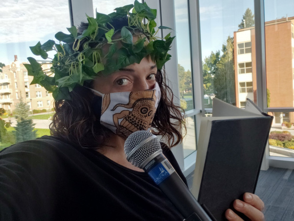 Woman with mask and garland on her head, holding book and speaking into microphone.