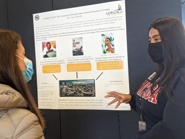 Poster showcasing student research in background as female student explains her work to another woman.