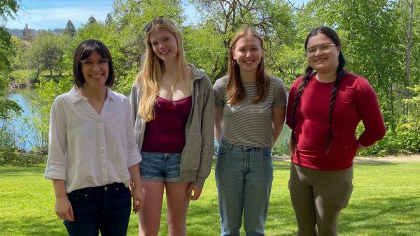 Four people (the Geck lab members) standing outside on a grassy college campus lawn on a sunny day